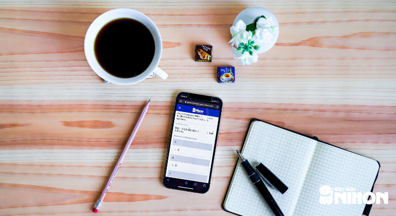 Foto tipo flat lay de uma xícara de café, dois chocolates, flores, um lápis, um smartphone com uma lição de japonês na tela, um caderno e uma caneta.