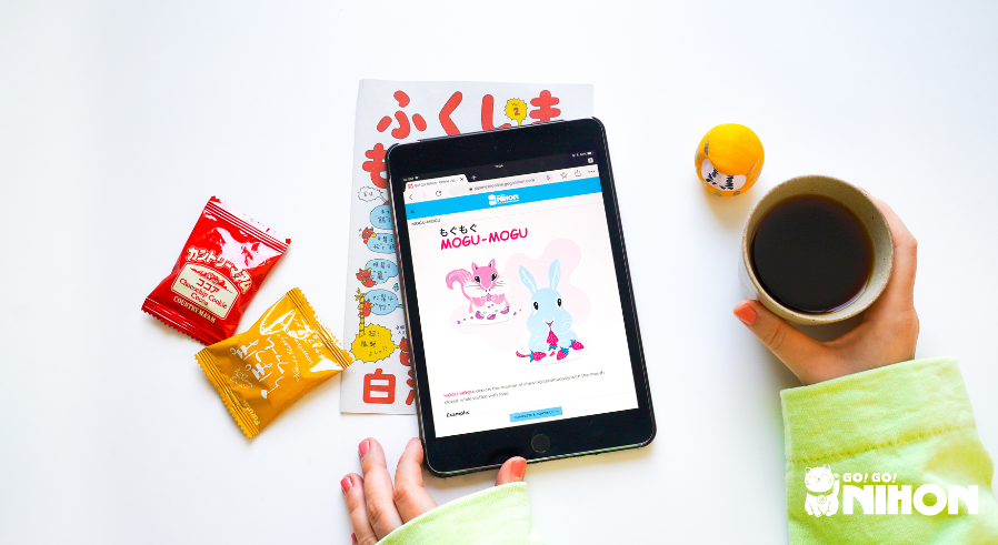 Eine Flatlay-Aufnahme eines Tablets mit einer Lektion zu Lautmalereien, daneben Snacks, ein Daruma und eine Tasse Kaffee