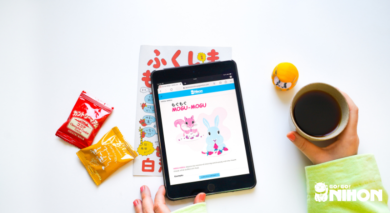 Imagen desde arriba de una tablet con una lección de japonés, galletas, daruma y una taza de té.