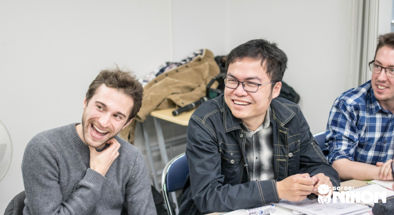 Tres estudiantes sonrientes sentados en escritorios en el aula aprendiendo japonés y divirtiéndose.