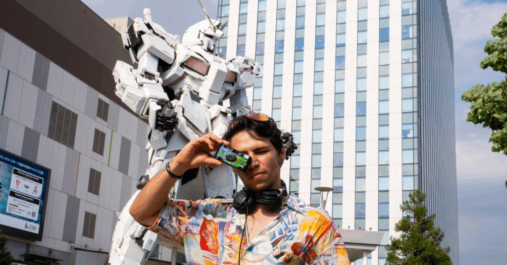 man holding up disposable camera in front of giant gundam statue
