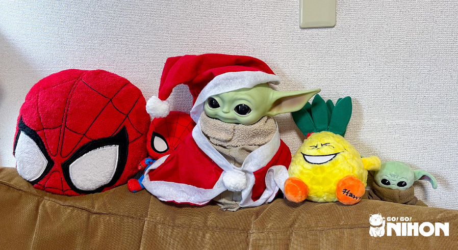 Baby Yoda dressed as Santa, Spider-man pillow, plushie, pineapple plushie, and mini Baby Yoda plushie sitting on top of a couch in a group