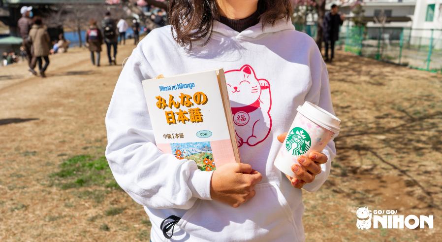 A woman in a Go! Go! Nihon sweatshirt holding a coffee cup and Japanese textbook.