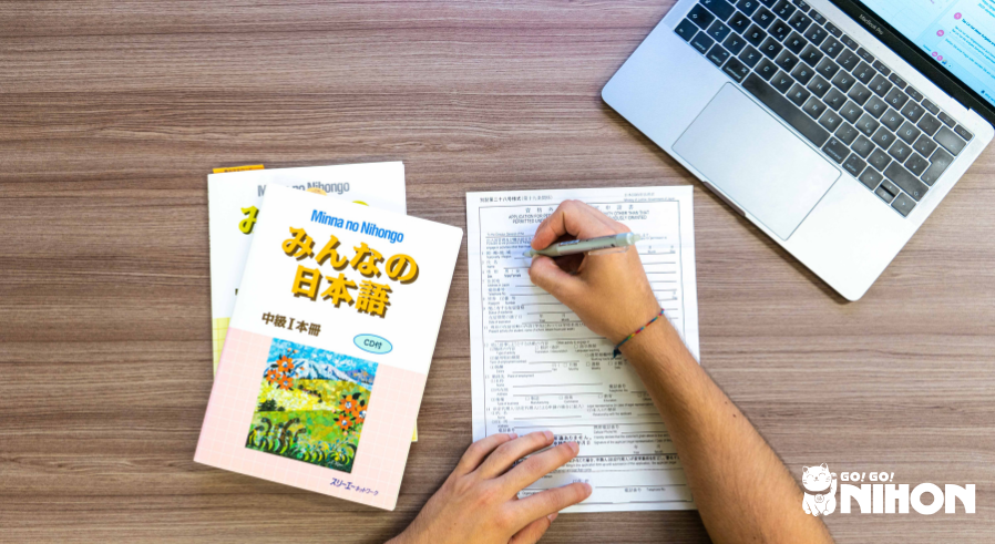 Flat lay photo of a person filling out a visa form on a desk with Japanese textbooks to the left and a laptop to the right