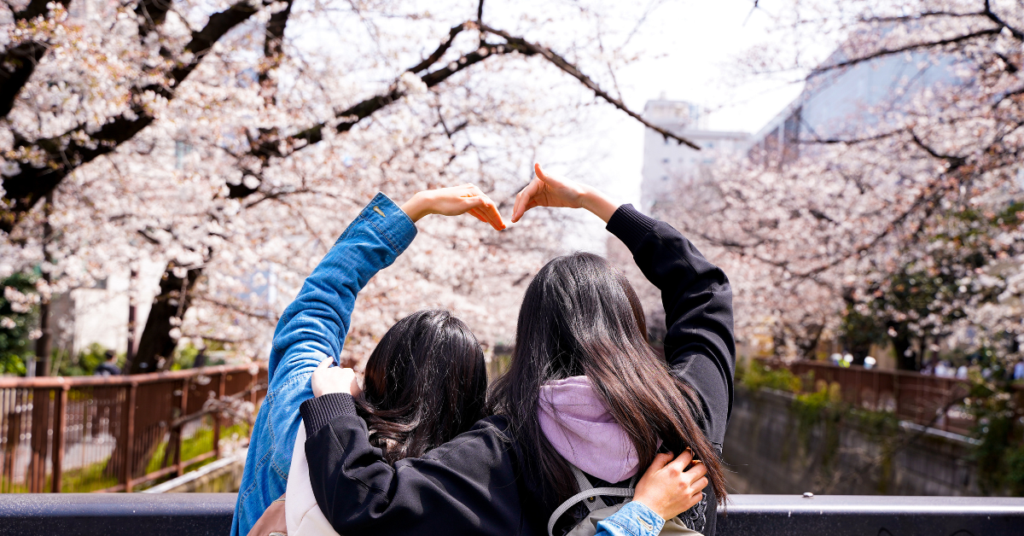 two women making a heart with their arms in front of sakura trees