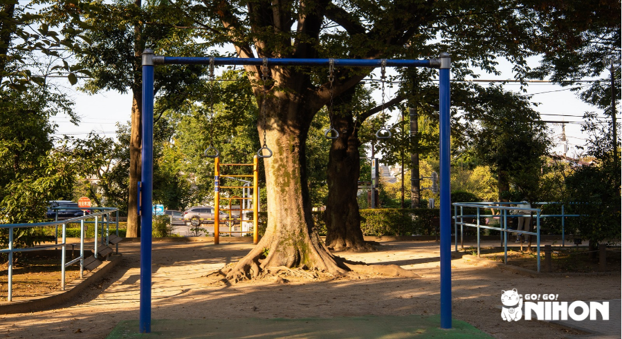 Struttura in metallo blu con anelli da ginnastica appesi in un parco giapponese ombreggiato, con un grande albero e un’attrezzatura per arrampicarsi arancione sullo sfondo